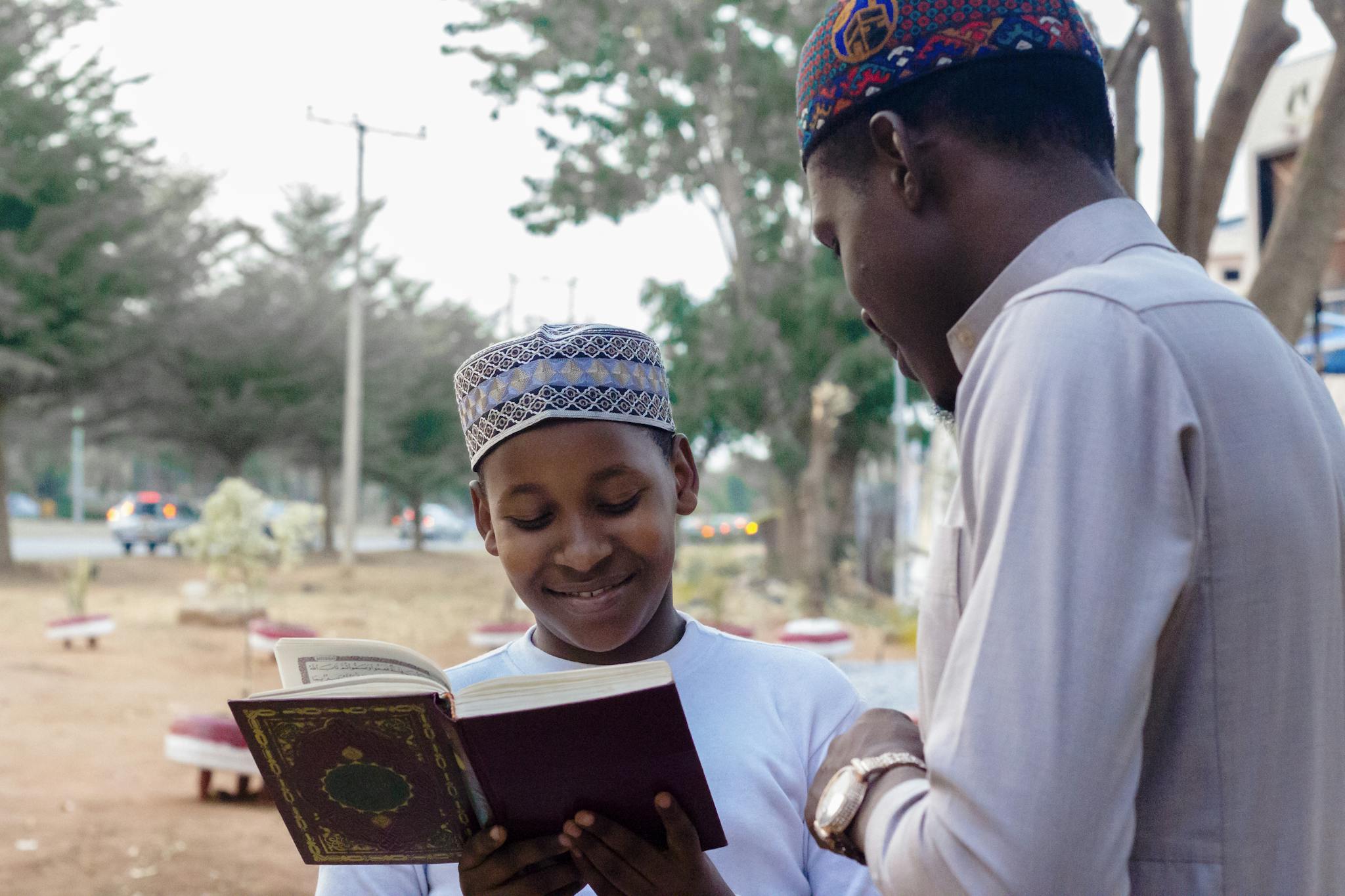 Young boy and man reading a religious book together while smiling, outdoors.